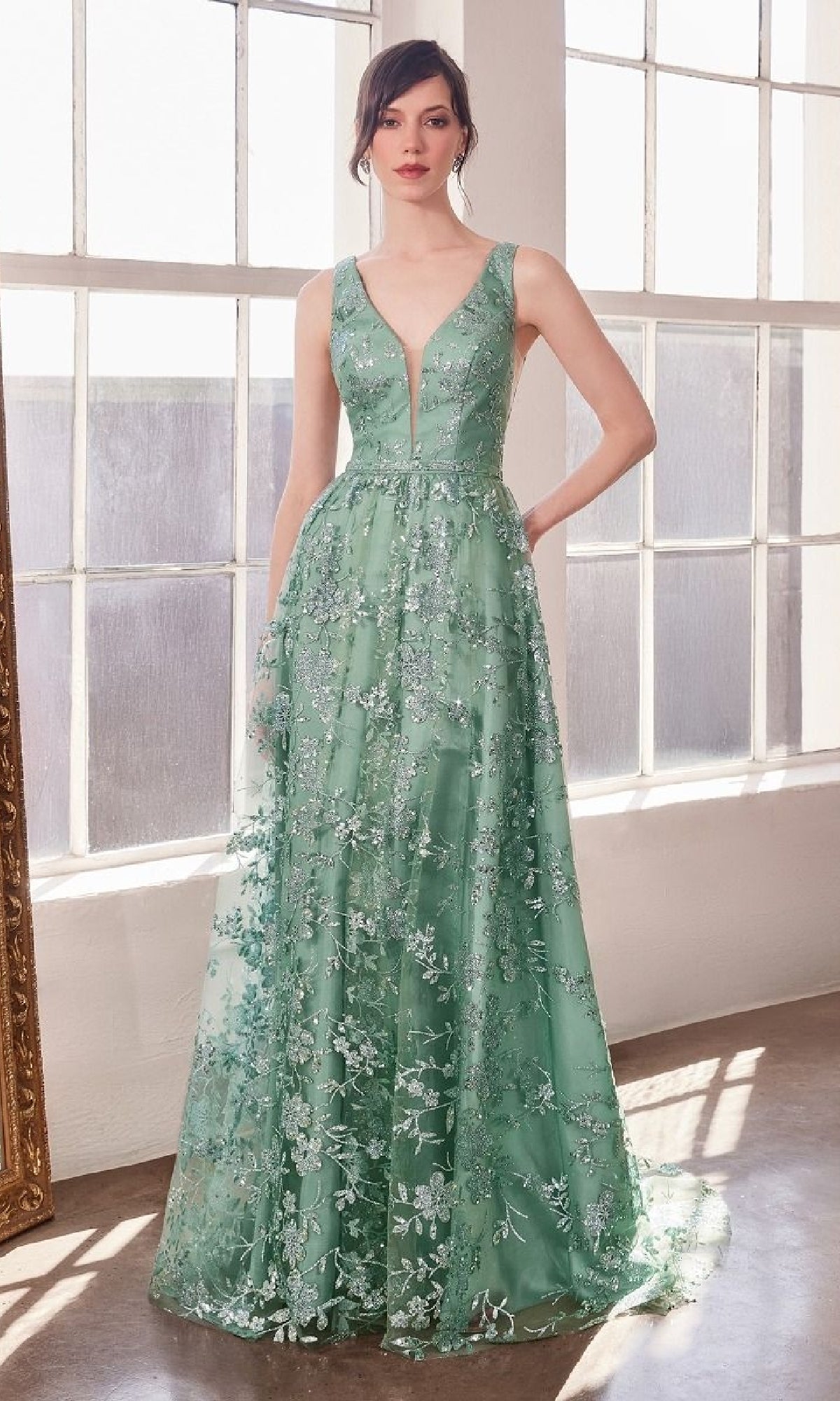 A woman stands indoors in the Amoris T606, an embroidered-lace long prom dress—floor-length, sleeveless, green with floral accents and a deep V-neckline.